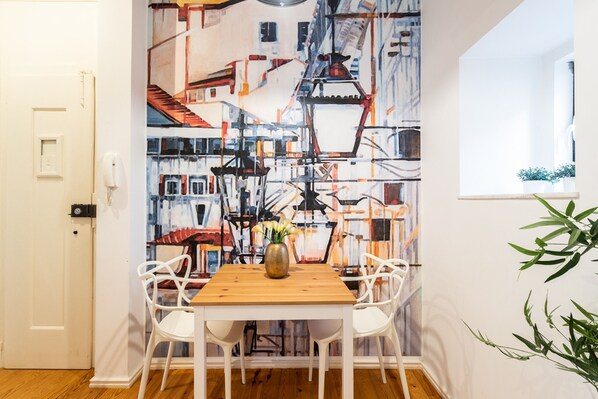 Zona pranzo con tavolo in legno e sedie bianche, un vaso con fiori e un quadro astratto colorato al muro.