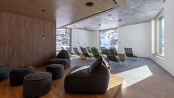 Un interno moderno con pavimento in legno, un'area relax con pouf e poggiapiedi, e vista su una montagna innevata.
