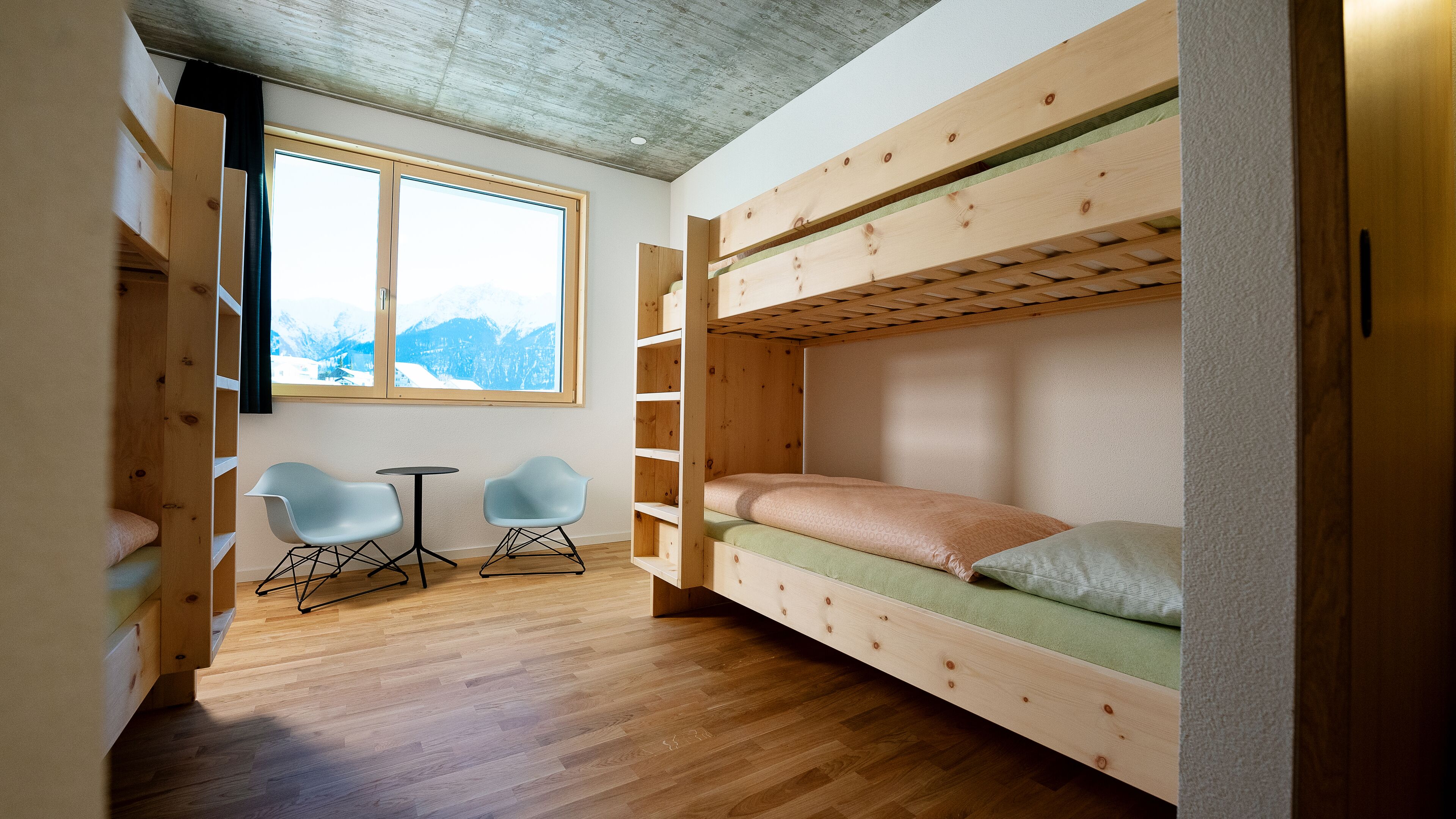 A cozy room with a bunk bed, two chairs, a small table, and a large window with a mountain view.
