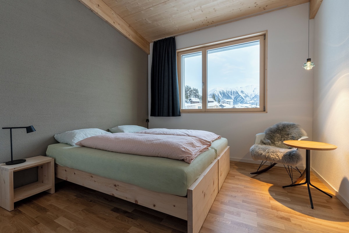 Quarto com cama de madeira, mesa de cabeceira, cadeira e uma pequena mesa.