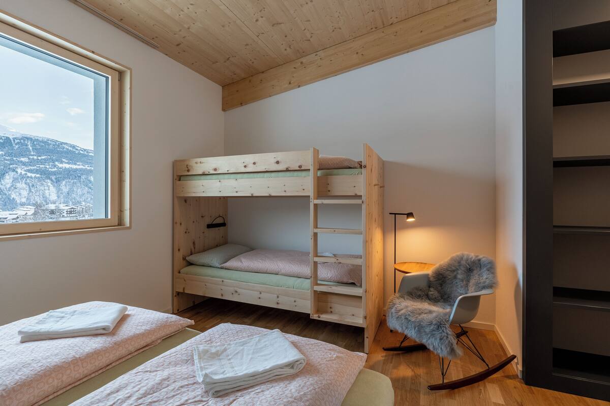 Quarto com cama beliche, cadeira de balanço e vista para uma paisagem nevada pela janela.
