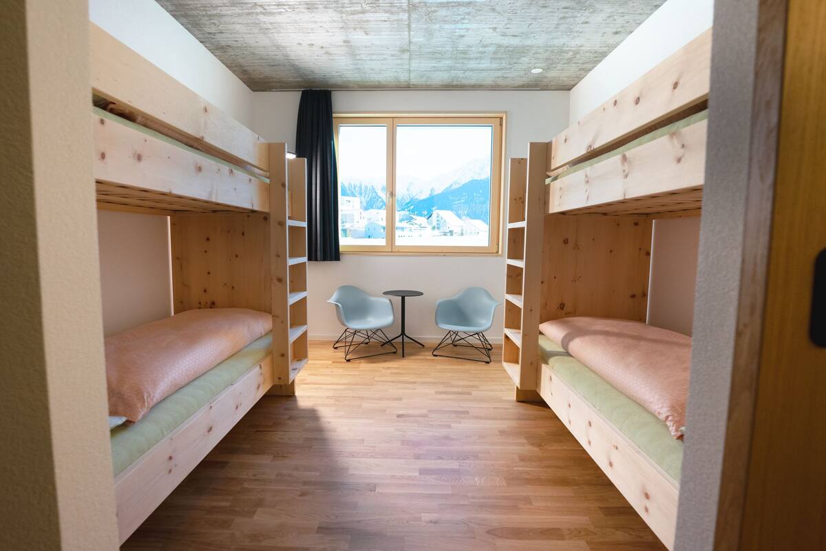 Quarto com duas beliches, uma mesa redonda pequena, duas cadeiras e uma janela com vista para as montanhas.