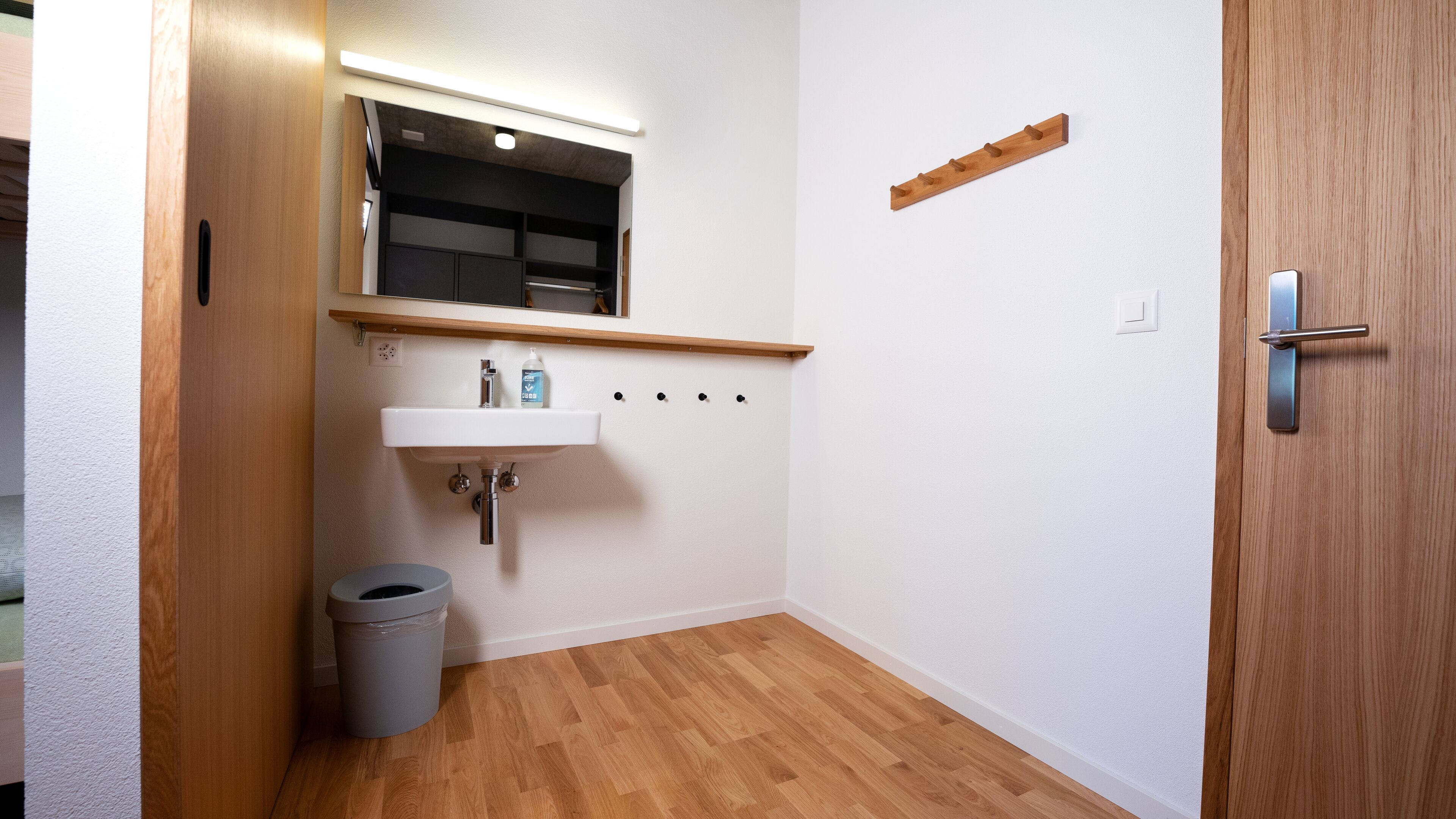 A small bathroom with a wooden floor, a white sink, a mirror above the sink, and a door with a handle.