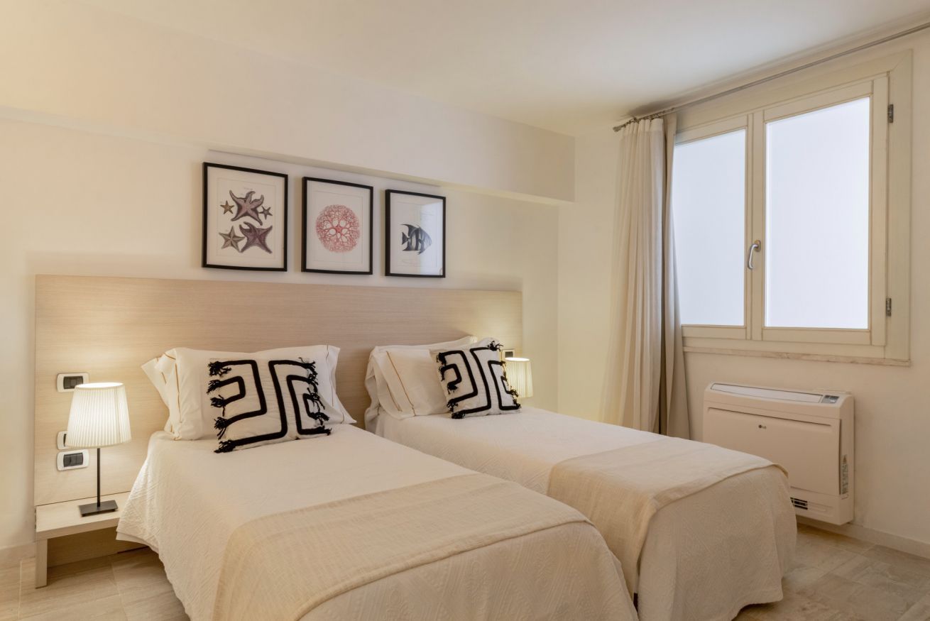 A hotel room with two beds, a nightstand with a lamp, a radiator, and framed artwork on the wall.