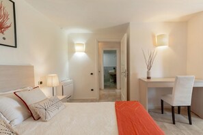 Habitación de hotel con cama, mesitas de noche, escritorio y silla. Se ve una puerta que da a un baño y un pasillo.