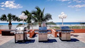 Three barbecue grills, a white fence, palm trees, and a view of the ocean. - The Seascape Premier Beachfront Cottage: Gulf View, Pool, Hot Tub, BBQ, WiFi (Indian Shores)