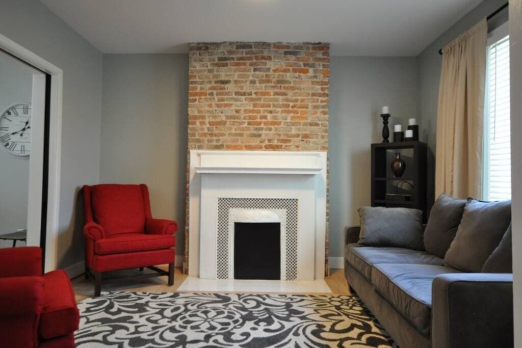 Un salon avec une cheminée en briques, un fauteuil rouge, un canapé gris, un tapis à motifs et une horloge au mur.