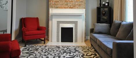 Un salon avec une cheminée en briques, un fauteuil rouge, un canapé gris, un tapis à motifs et une horloge au mur.
