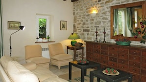 Un salon chaleureux avec un canapé, deux fauteuils, une table basse et un meuble en bois. On y trouve un mur en pierre, une fenêtre avec des rideaux et une lampe sur une table d’appoint.
