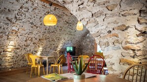 Uma área de jantar rústica com parede de pedra, mesa de madeira, cadeiras amarelas e vermelhas, luminárias pendentes e um vaso com planta.