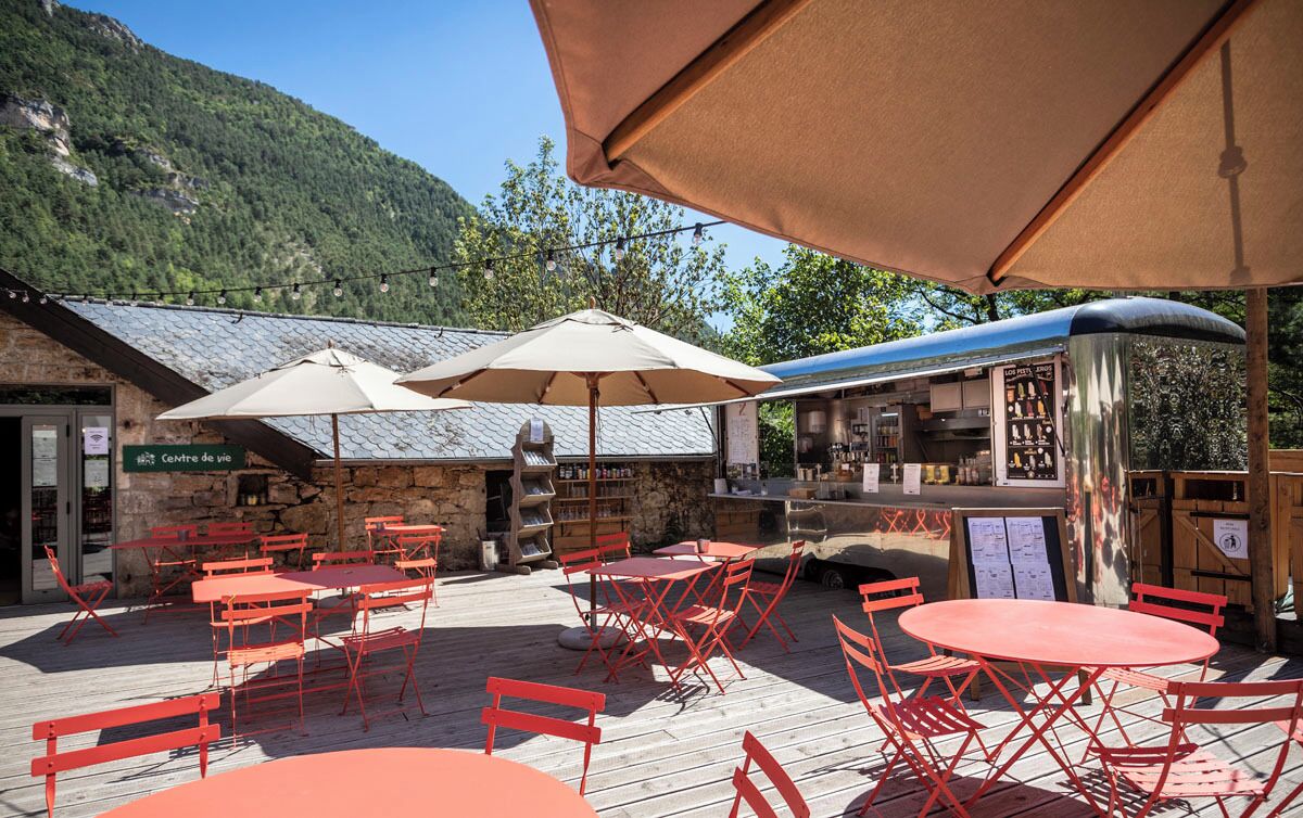 An outdoor café with red metal tables and chairs, beige umbrellas, and a stone building with a sign reading "Centre de vie".