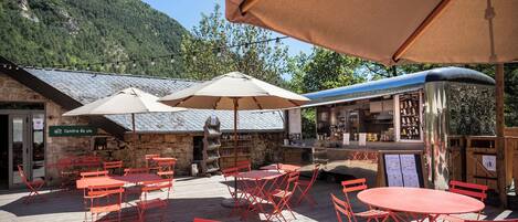 An outdoor café with red metal tables and chairs, beige umbrellas, and a stone building with a sign reading "Centre de vie".