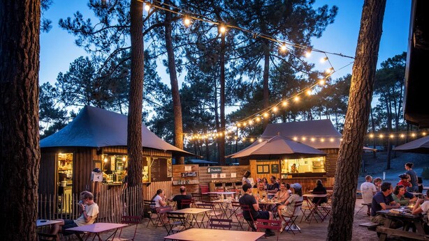 An outdoor dining area with tables and chairs, surrounded by trees and string lights. - Huttopia Lac de Carcans (Carcans)