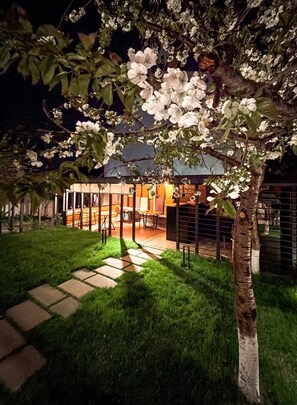 A well-lit outdoor seating area with a dining table and chairs, surrounded by a fence and a tree with white blossoms. - Andor Hotel (Yerevan)