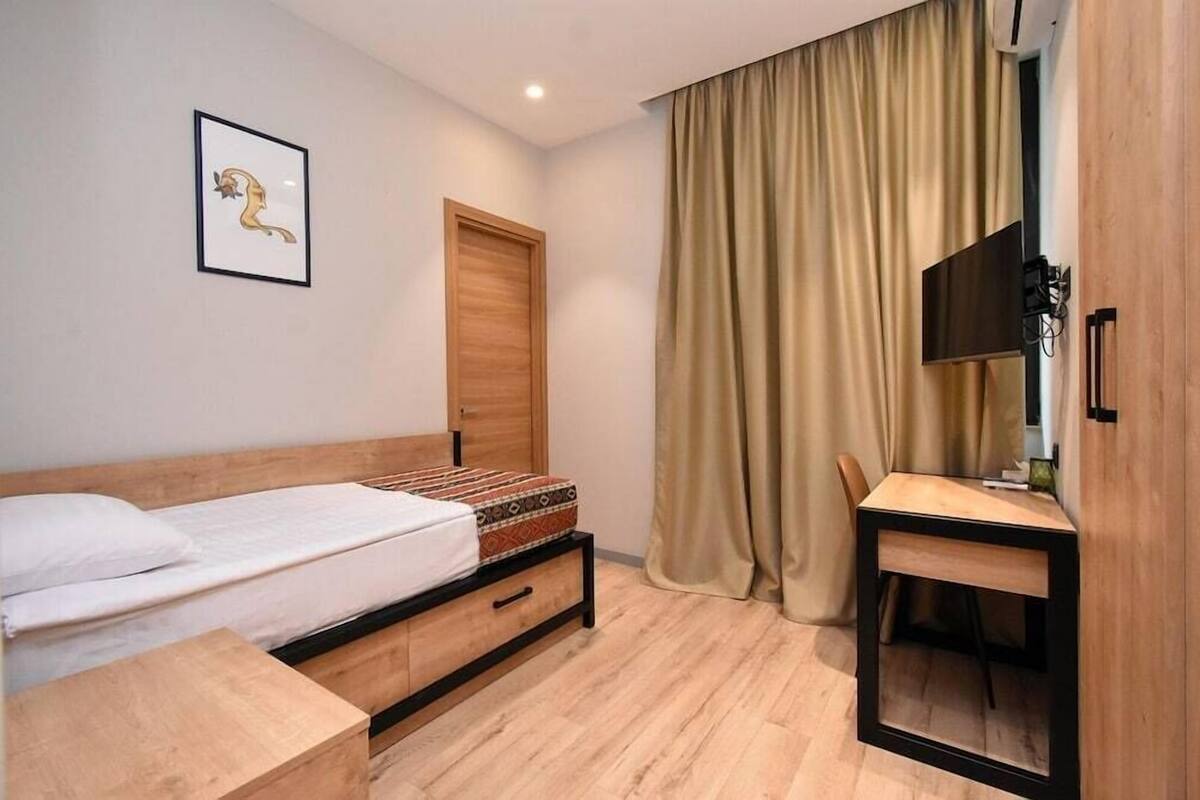 A bedroom with a bed, a desk, a chair, a wooden wardrobe, a framed picture on the wall, and a window with curtains.