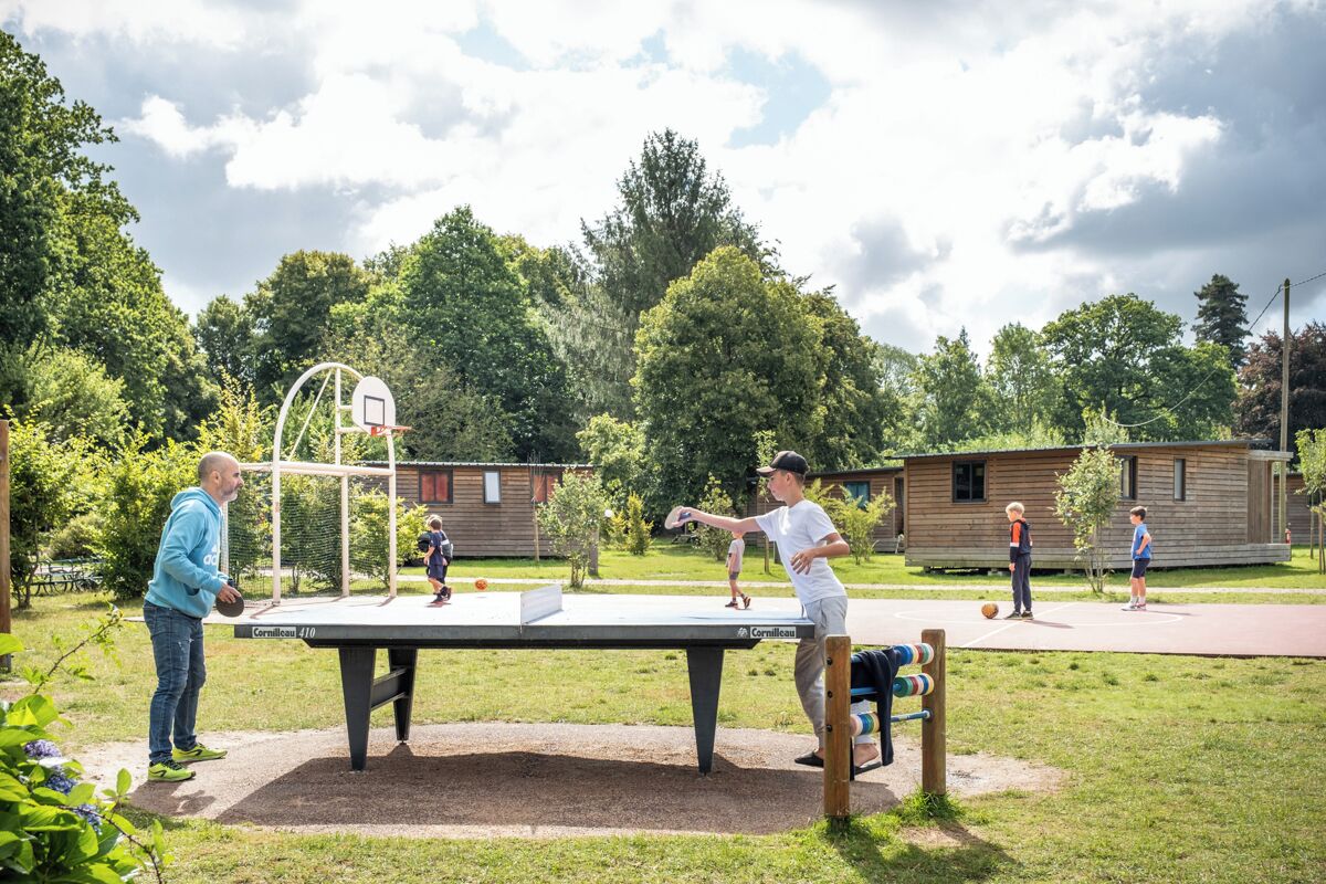 Een tafeltennistafel in een buitenomgeving, met een basketbalveld en een houten gebouw op de achtergrond.