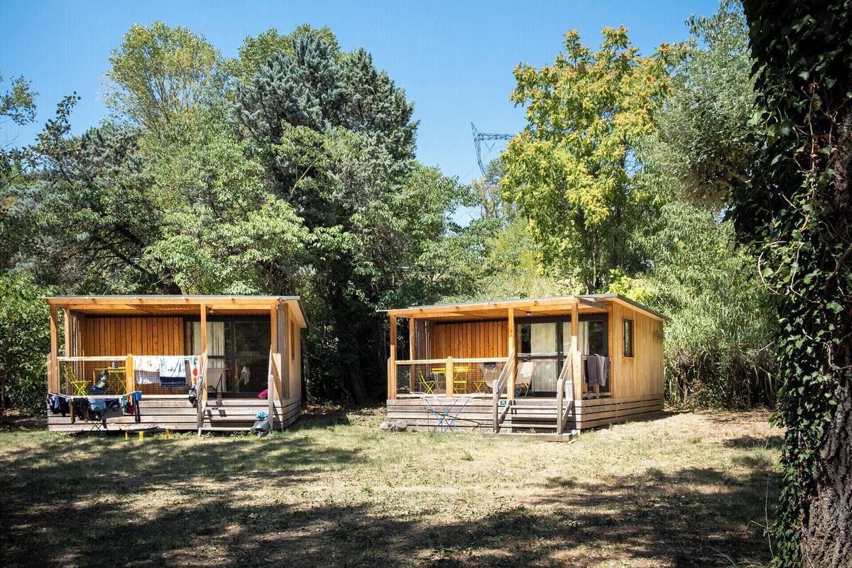 Two wooden cabins with balconies, surrounded by dense trees and a clear sky.