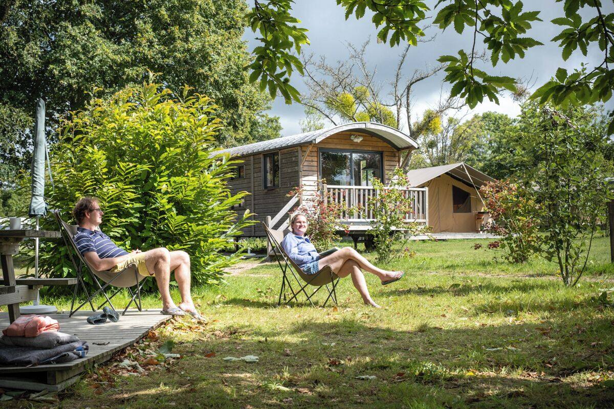 Two people relaxing in lawn chairs in a grassy area with a wooden deck and a small house in the background.