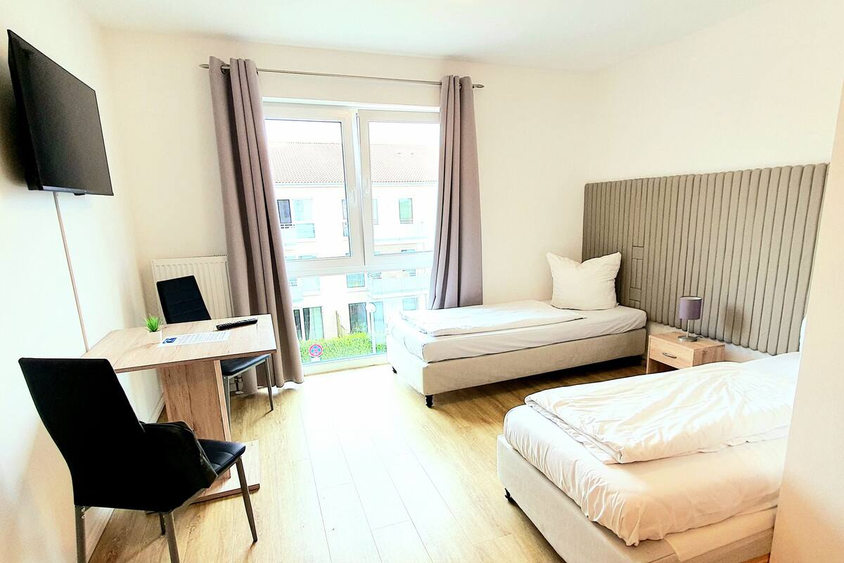 A hotel room with a bed, a desk with a chair, a TV mounted on the wall, and a window with curtains.
