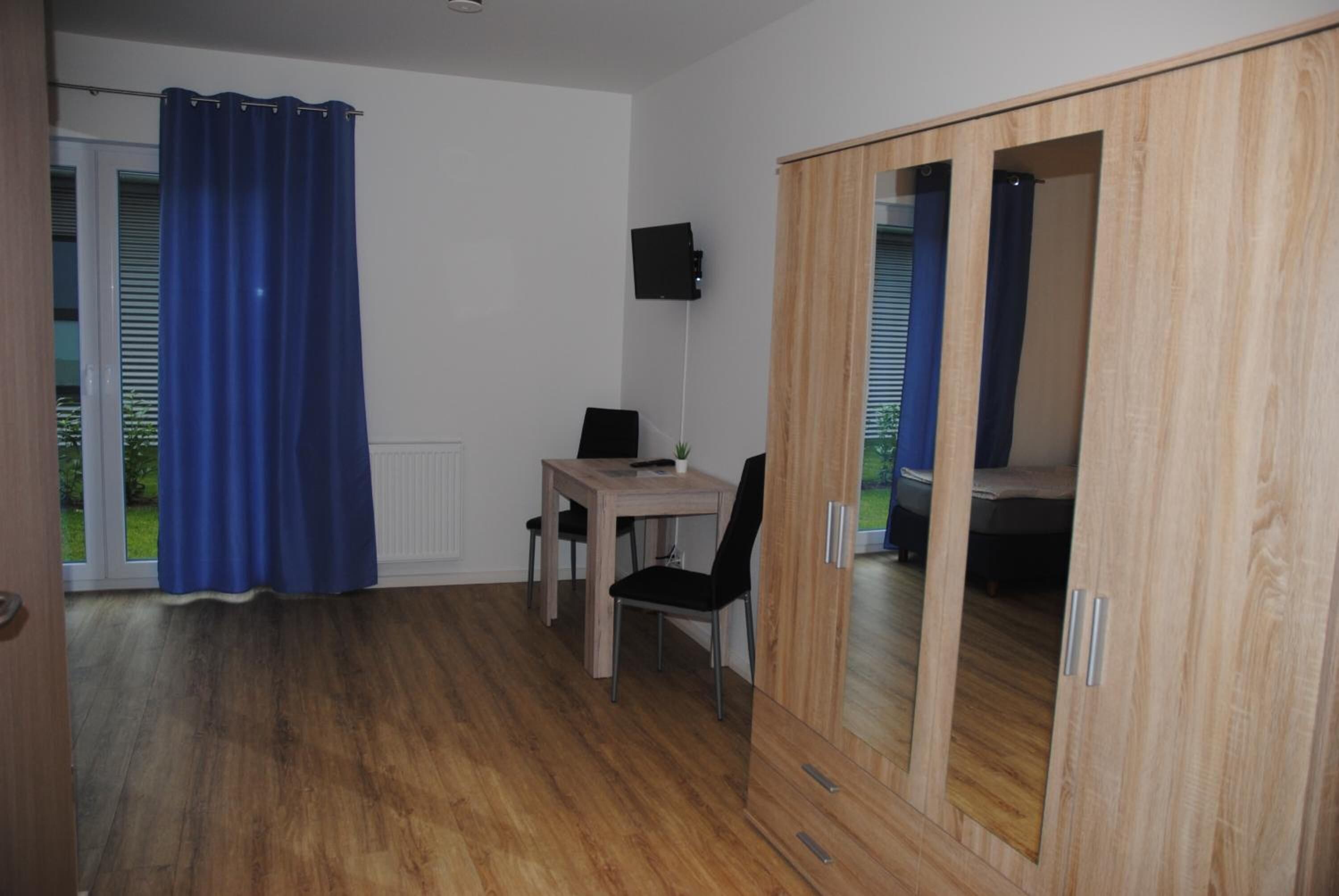 A room with a wooden floor, a table with a chair, a blue curtain, and a wardrobe with sliding doors.