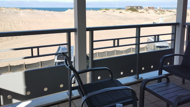 Ein Balkon mit Blick auf einen Sandstrand und das Meer. Zwei Stühle und ein Tisch sind vorhanden.