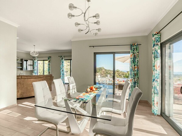 Un comedor moderno con una mesa de vidrio y sillas blancas, una cocina al fondo y un amplio ventanal con vistas al exterior.