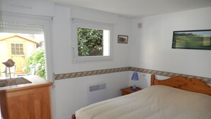 Un dormitorio con una cama de madera, una mesita de noche con lámpara, una ventana con vista a la vegetación y un cuadro pequeño en la pared.