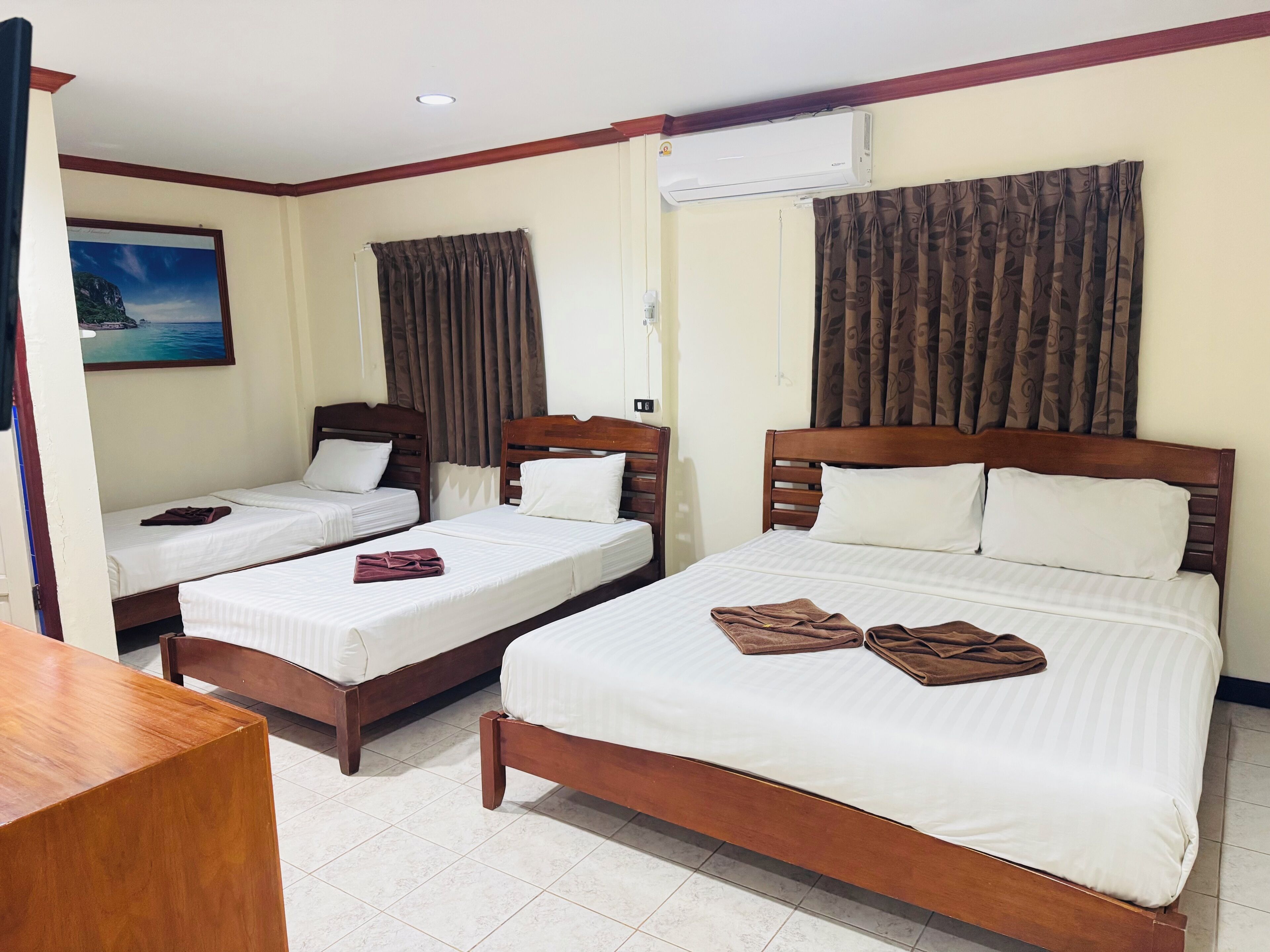 A hotel room with two single beds and one double bed, wooden headboards, white linens, brown towels, a painting of a coastal scene, and an air conditioning unit.