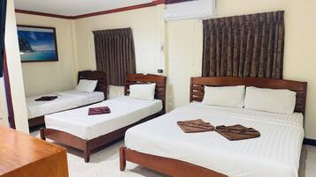 A hotel room with two single beds and one double bed, wooden headboards, white linens, brown towels, a painting of a coastal scene, and an air conditioning unit.