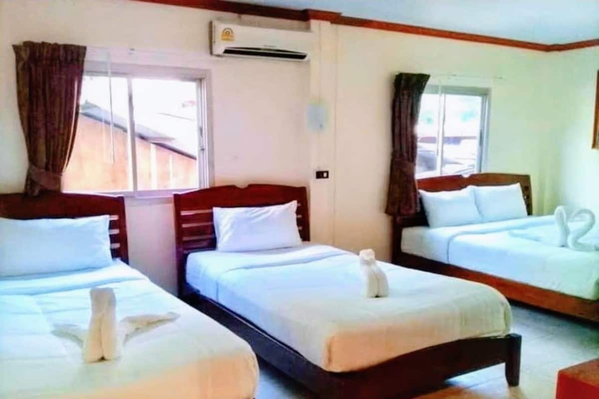 A hotel room with three beds, each with white linens and a towel on the beds. There is an air conditioning unit on the wall above the window.