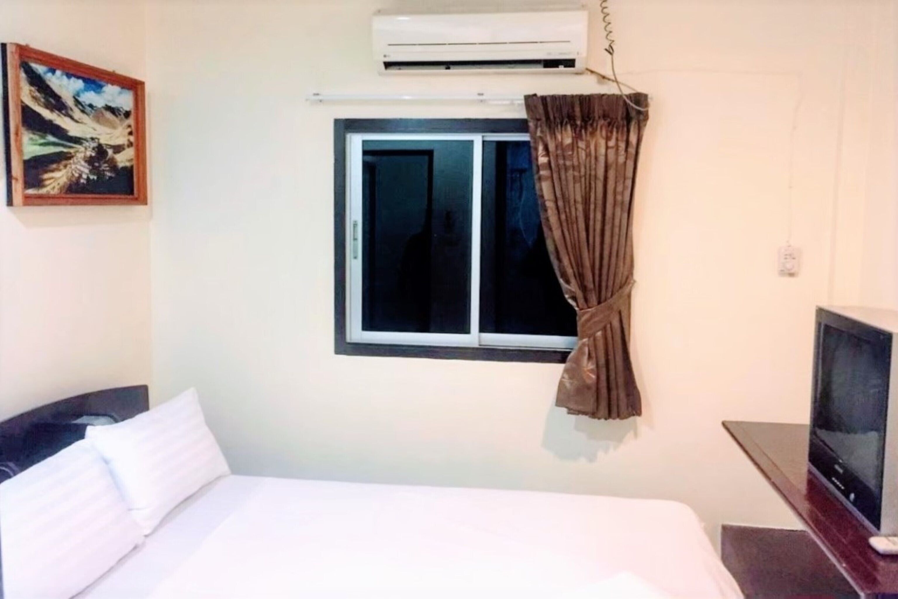 A hotel room with a bed, a television, an air conditioning unit, a window with curtains, and a painting on the wall.