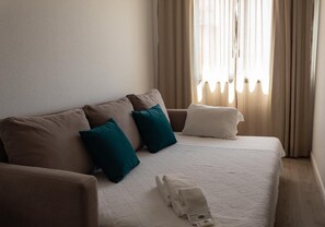 A neatly made bed with a white comforter and two pillows, a brown sofa with teal and white pillows, and a window with sheer curtains allowing natural light. - Hotel Santa Cecília (Ourem)