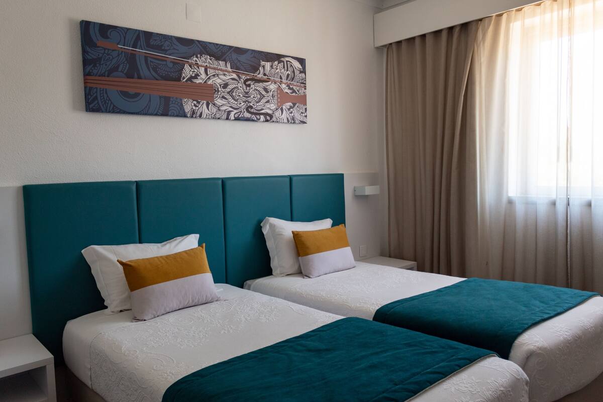 Hotellihuone, jossa on kaksi sänkyä, turkoosinsiniset päätyosat, valkoiset vuodevaatteet ja suuri abstrakti maalaus seinällä.