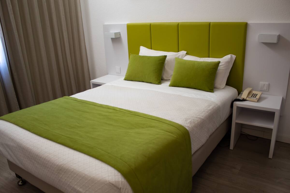 Hotellihuone, jossa on sänky, vihreät ja valkoiset vuodevaatteet, vihreä sängynpääty, valkoiset yöpöydät ja puhelin toisella yöpöydällä.