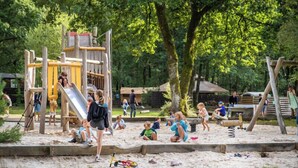 A playground with a slide and climbing structures, surrounded by sand and trees.
