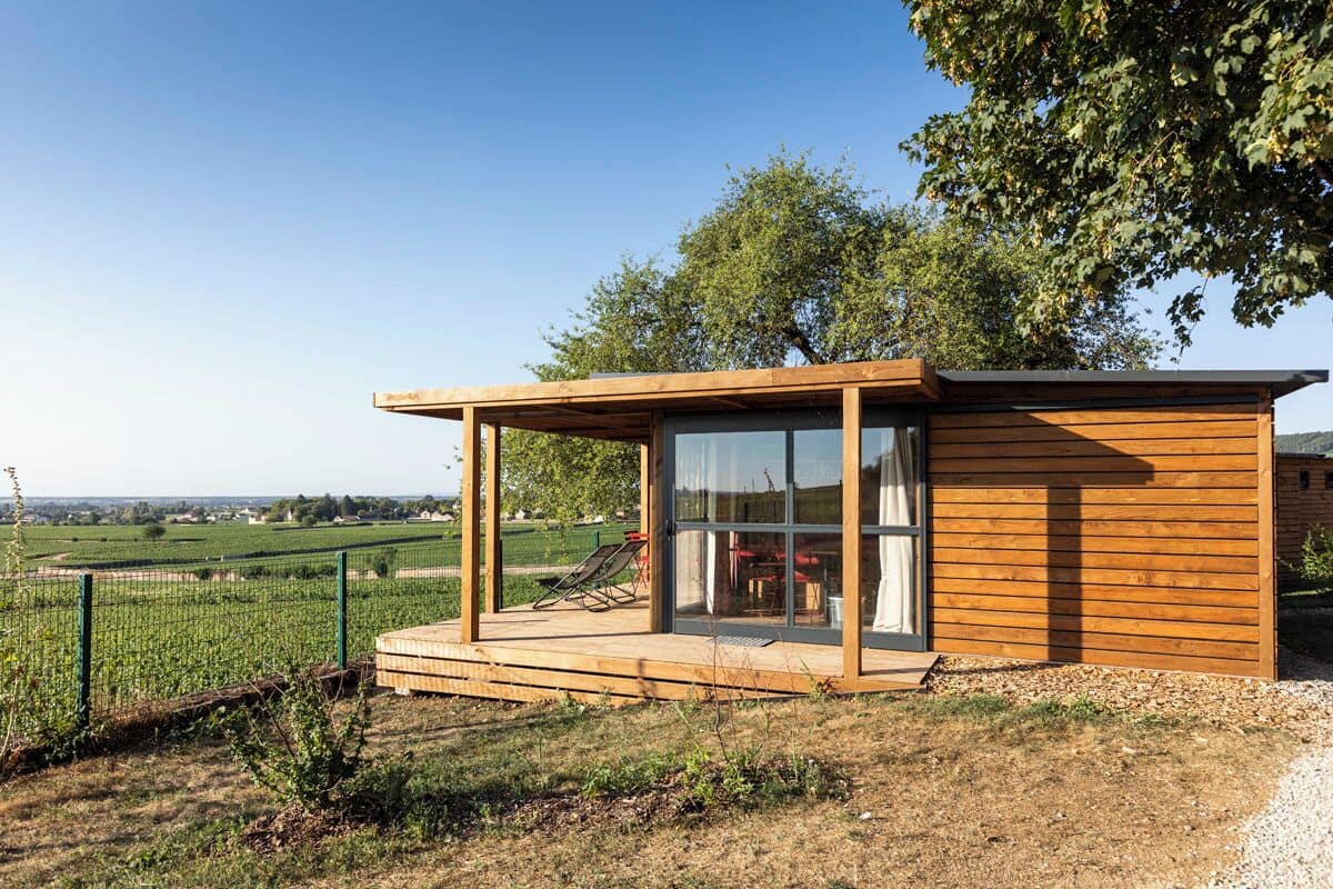 Une cabane moderne en bois, dotée d’une porte en verre, d’une terrasse et offrant une vue sur un vignoble.