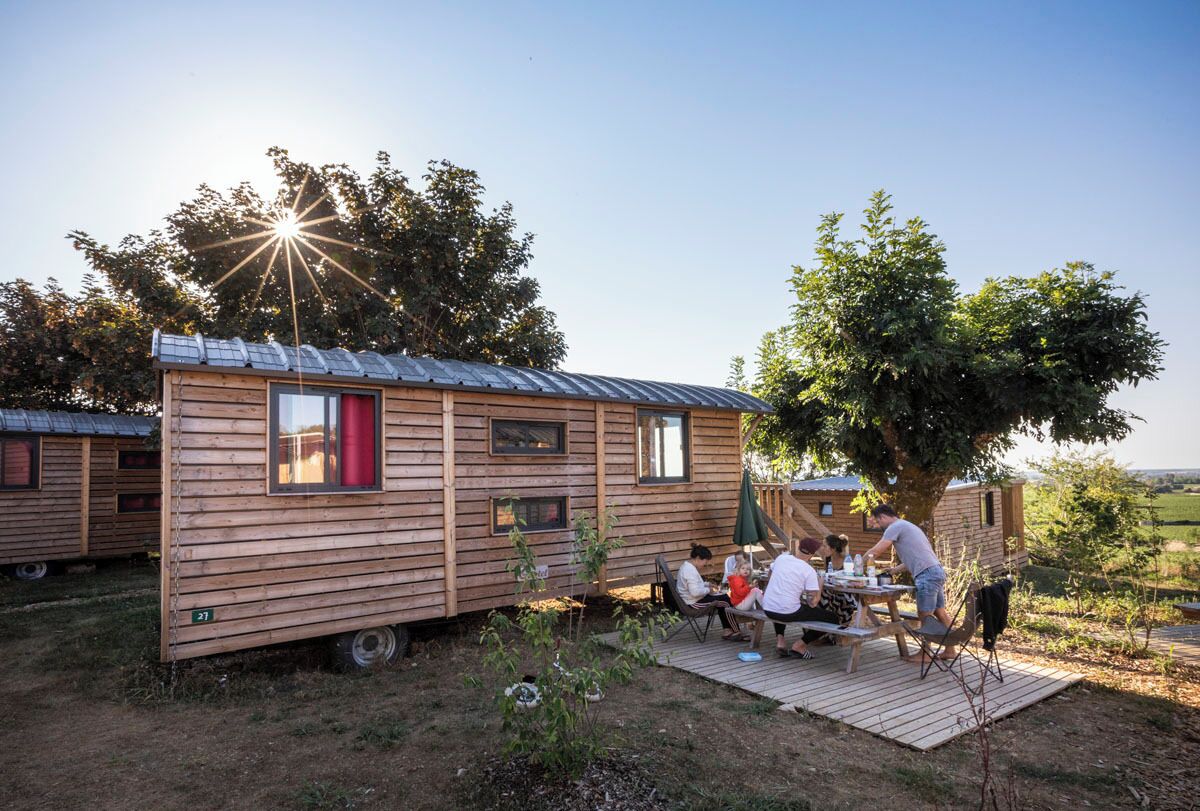 A wooden cabin with a metal roof, a dining area with a table and chairs, and a tree in the background.