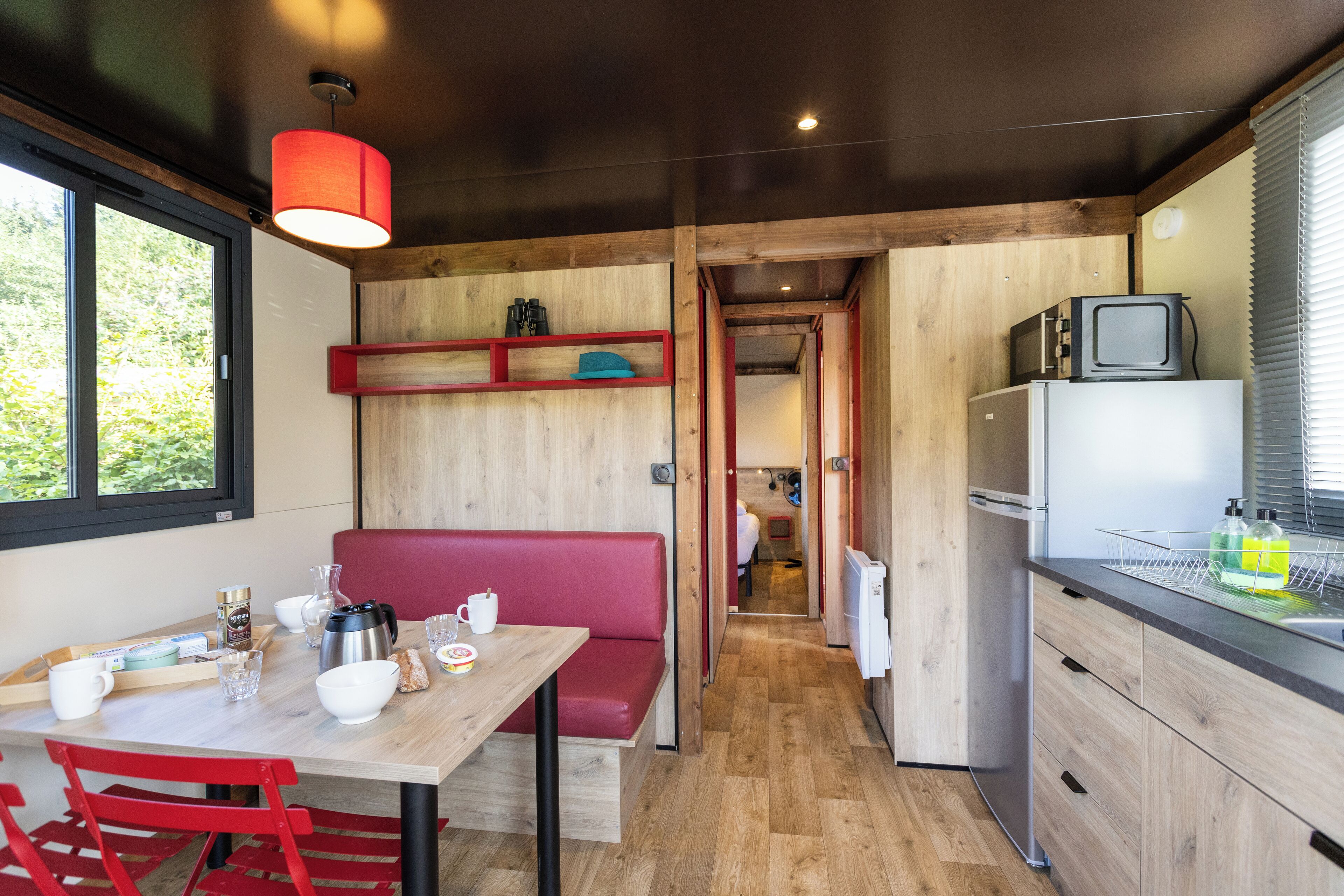 A compact kitchen with wooden cabinets, a dining table with red chairs, a small refrigerator, and a television on top.