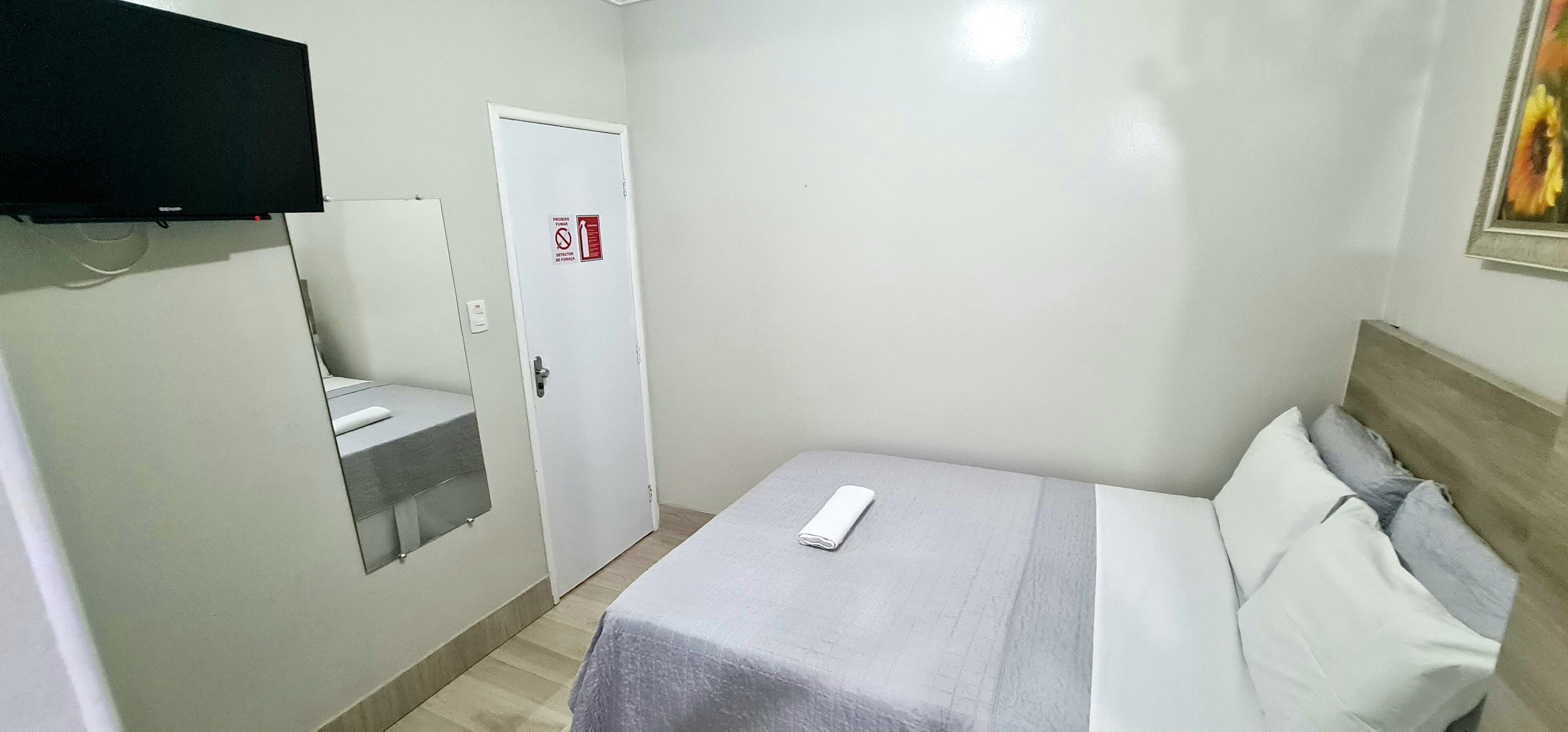 A hotel room with a bed, a TV mounted on the wall, a door with a no-smoking sign, and a painting on the wall.