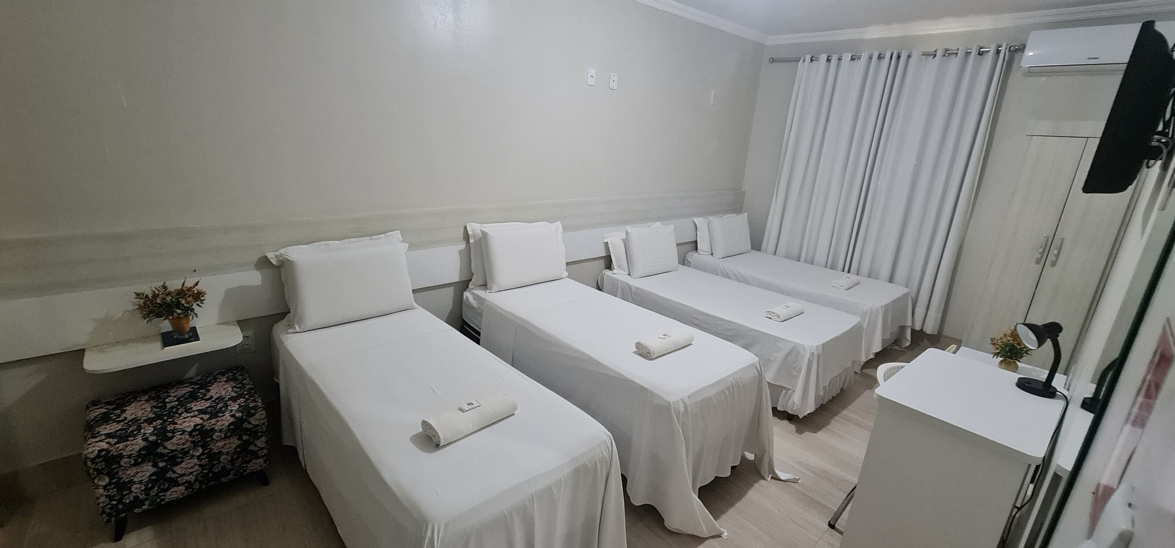 A hotel room with four single beds, a small desk with a lamp, a potted plant, and a suitcase.