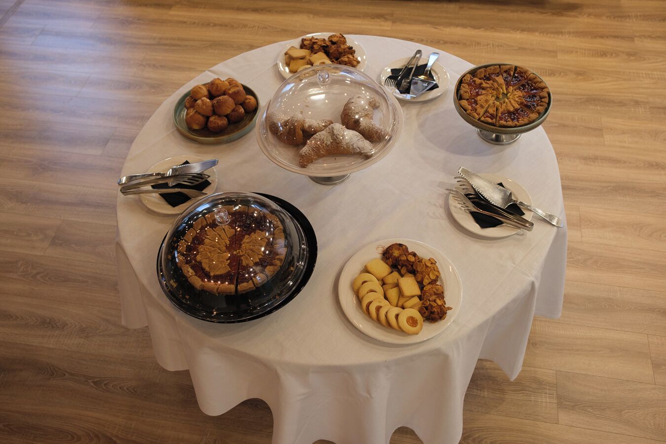 Una mesa redonda con diversos postres y pasteles, incluyendo un pastel de frutas, hojaldras y un bizcocho, sobre un mantel blanco.