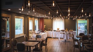 A dining area with multiple tables, hanging light fixtures, and large windows providing natural light. - Lake Hotel La Pieve (Pisogne)