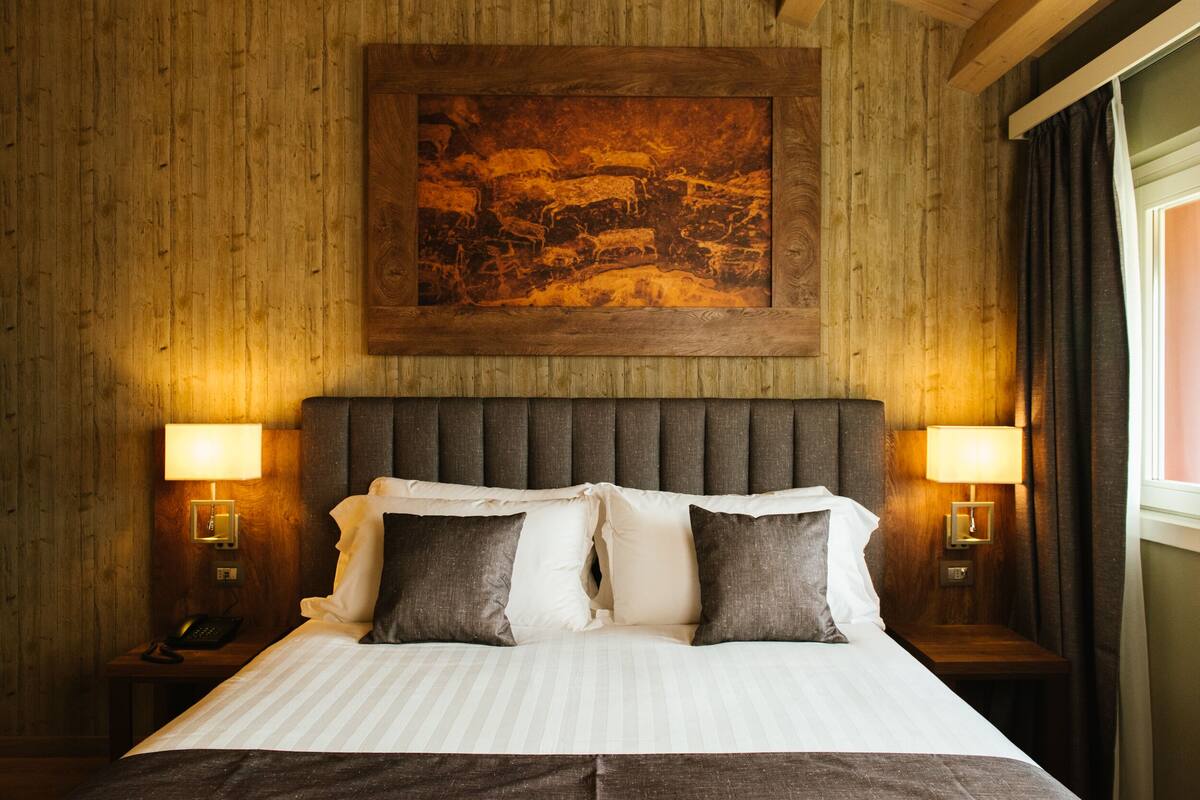 A hotel room with a wooden headboard, two bedside lamps, a large framed artwork above the bed, and a window with curtains.