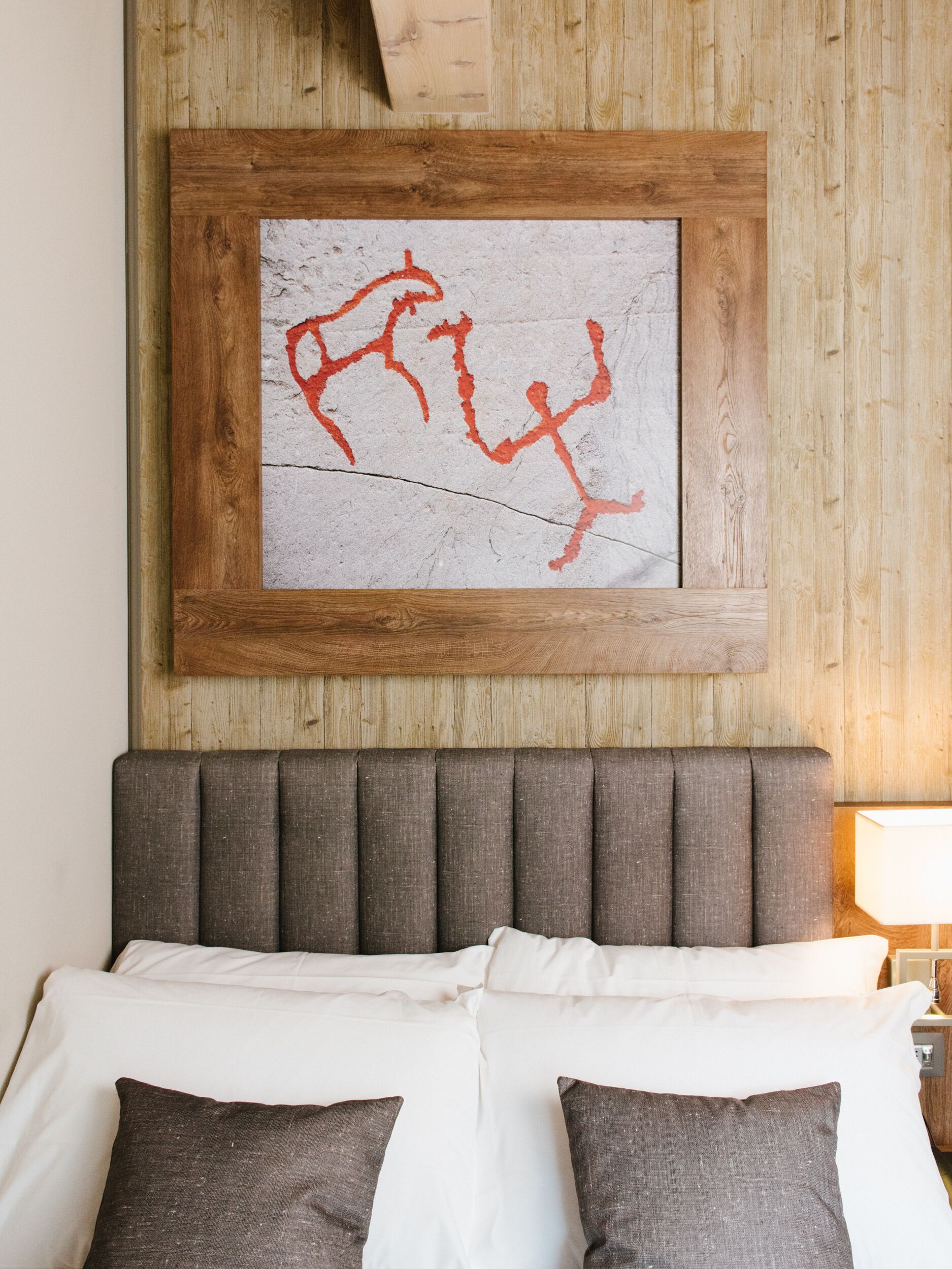 Ein Bett mit Holz-Kopfseitenverkleidung, weißer Bettwäsche und zwei dunklen Kissen. Über dem Bett hängt ein gerahmtes Kunstwerk mit roter Schrift.