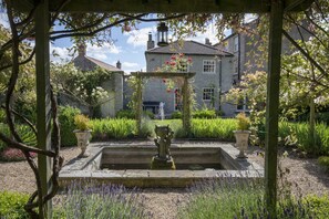 A garden with a fountain, a pergola, and a stone building in the background.