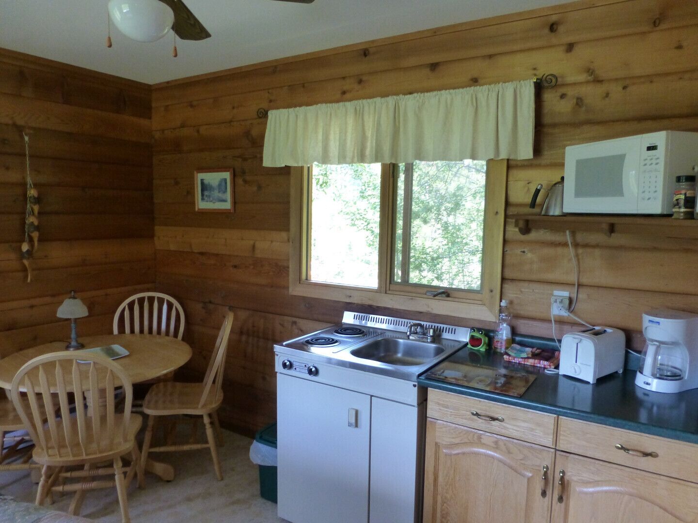 A compact kitchen with wooden walls, a dining table with chairs, a stove, a microwave, and a window with a view of greenery.