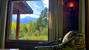 Une pièce avec vue sur une forêt et des montagnes, visible par une fenêtre, comprenant un fauteuil à motifs et une lampe décorative.