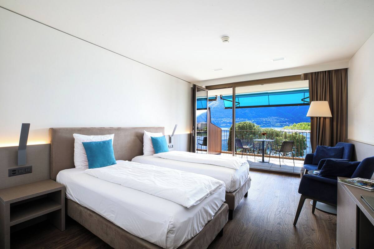 A modern hotel room with two beds, a desk, and a balcony with a view of the sea.