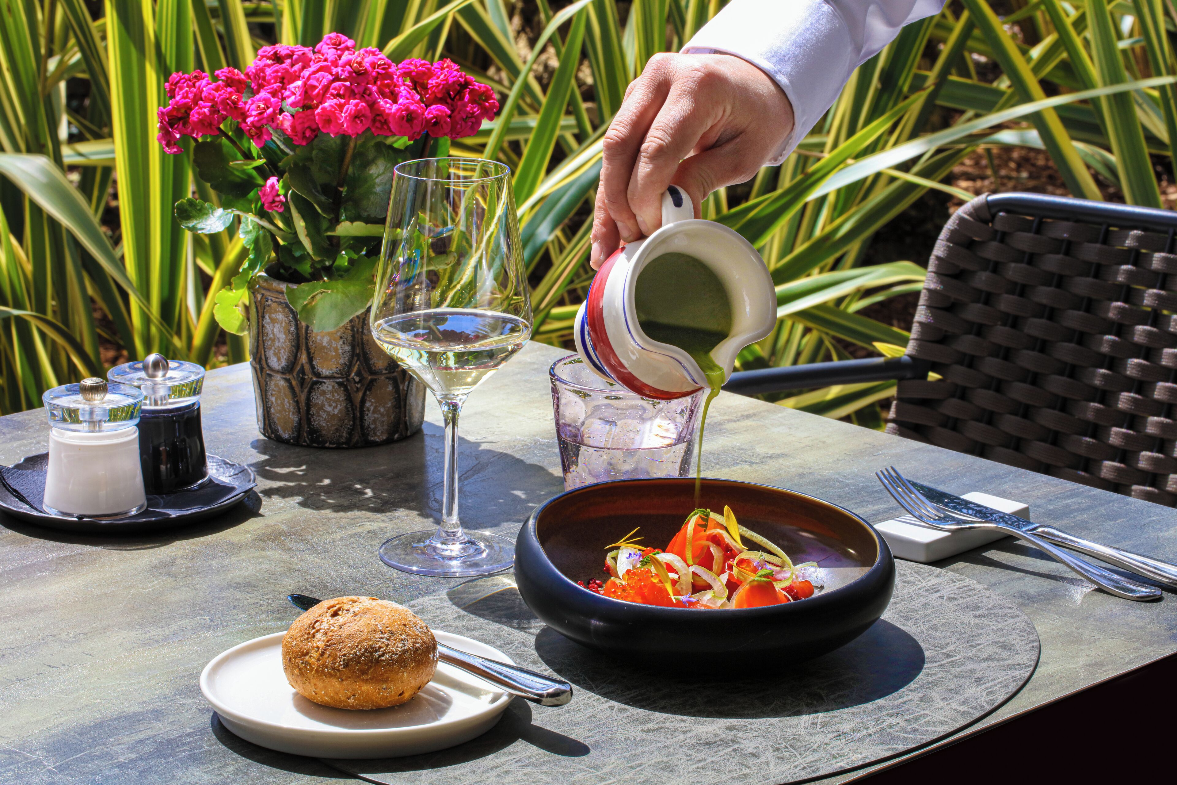 A table set with a bowl of food, a glass of white wine, a glass of water, a small plate with a pastry, and a vase with pink flowers.
