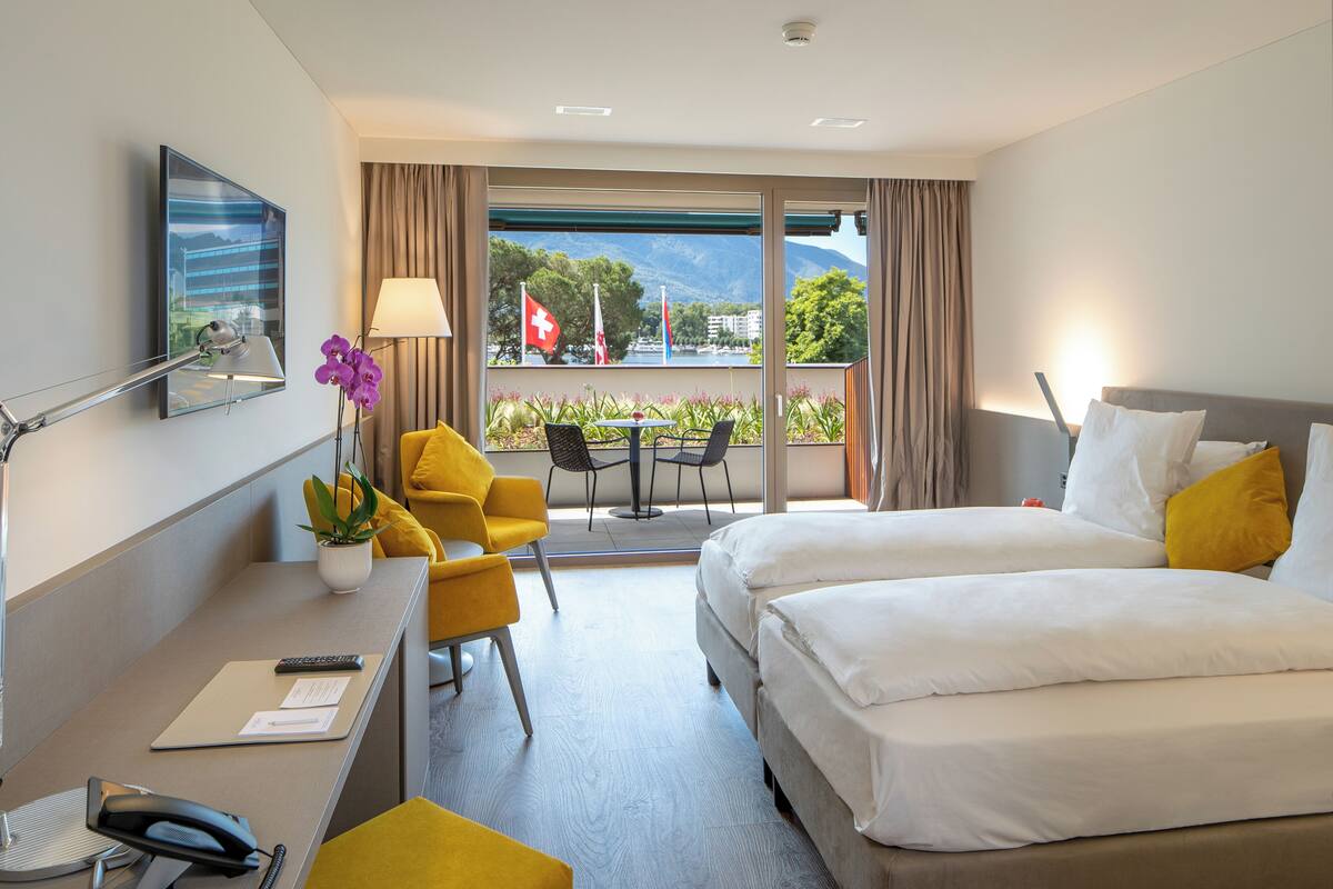 A hotel room with a bed, desk, chair, and a balcony with a view of the mountains and lake.